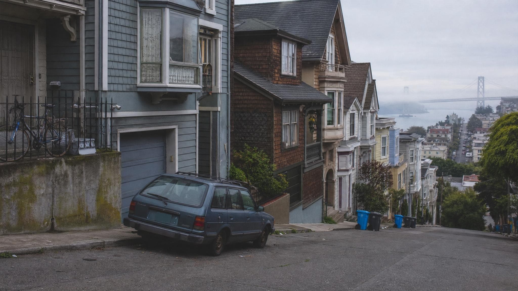 A steep San Francisco hillside street on a foggy morning, lined with Victorian and wood-shingle homes, with San Francisco Bay and the Bay Bridge in the distance.
