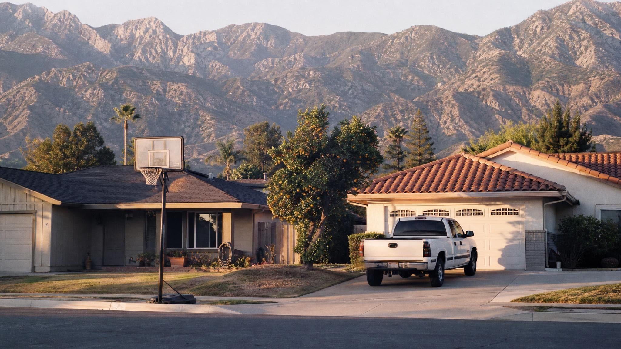 An Inland Empire ranch-home street in the Rancho Cucamonga area at golden hour, with a citrus tree and the San Bernardino mountains rising behind the rooftops.
