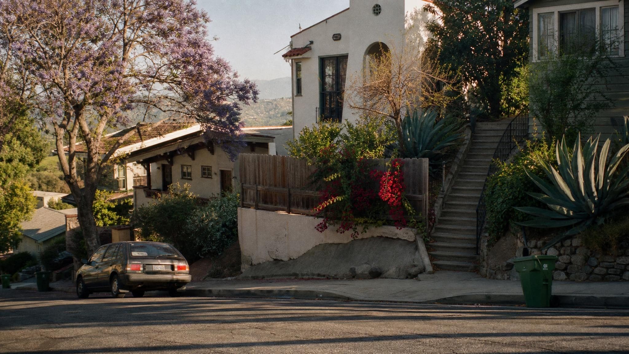 A Silver Lake hillside street in Los Angeles at golden hour, with a Spanish Revival home, a blooming jacaranda, a public stairway, and the San Gabriel foothills beyond.