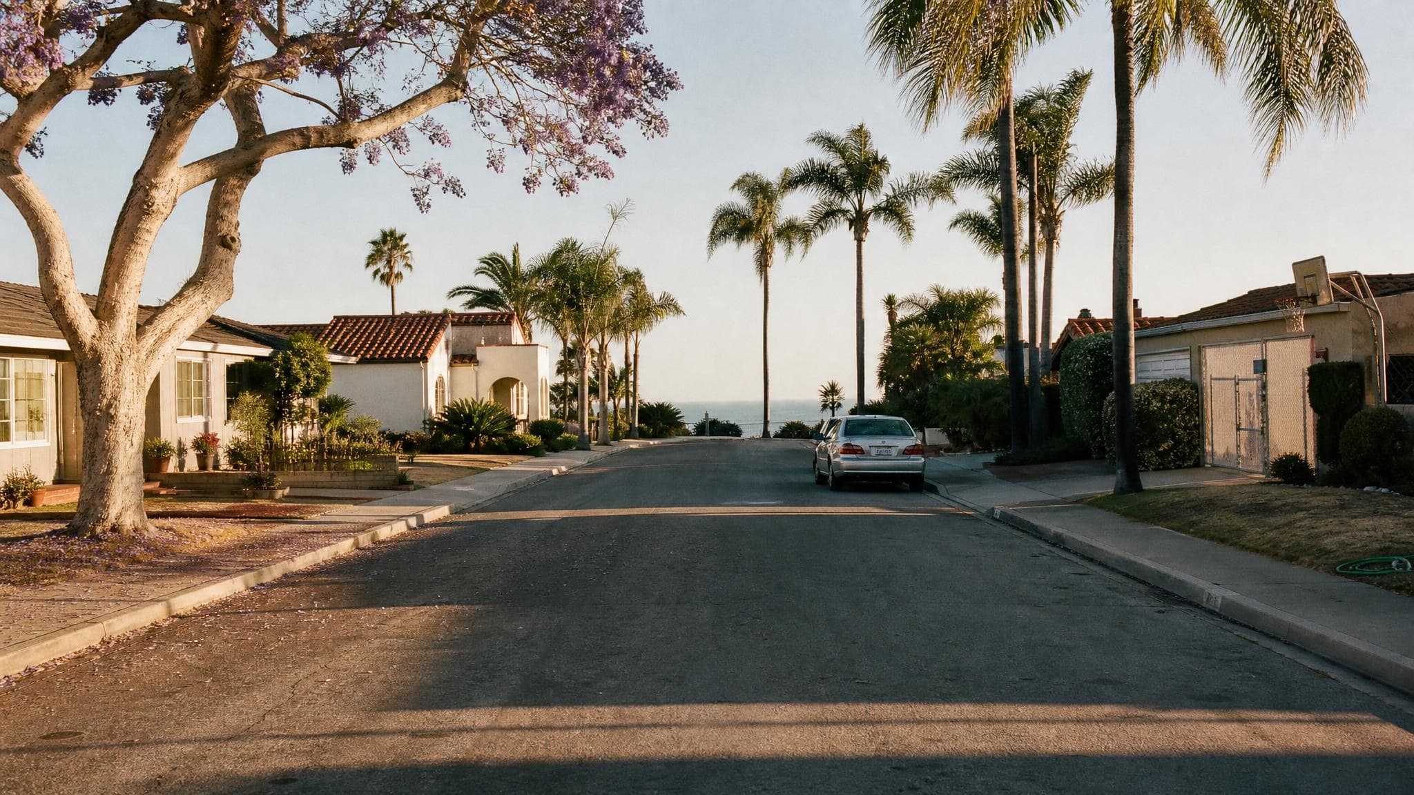 A quiet coastal Orange County residential street at golden hour, lined with palms and Spanish-style homes, with the Pacific visible at the end of the block.