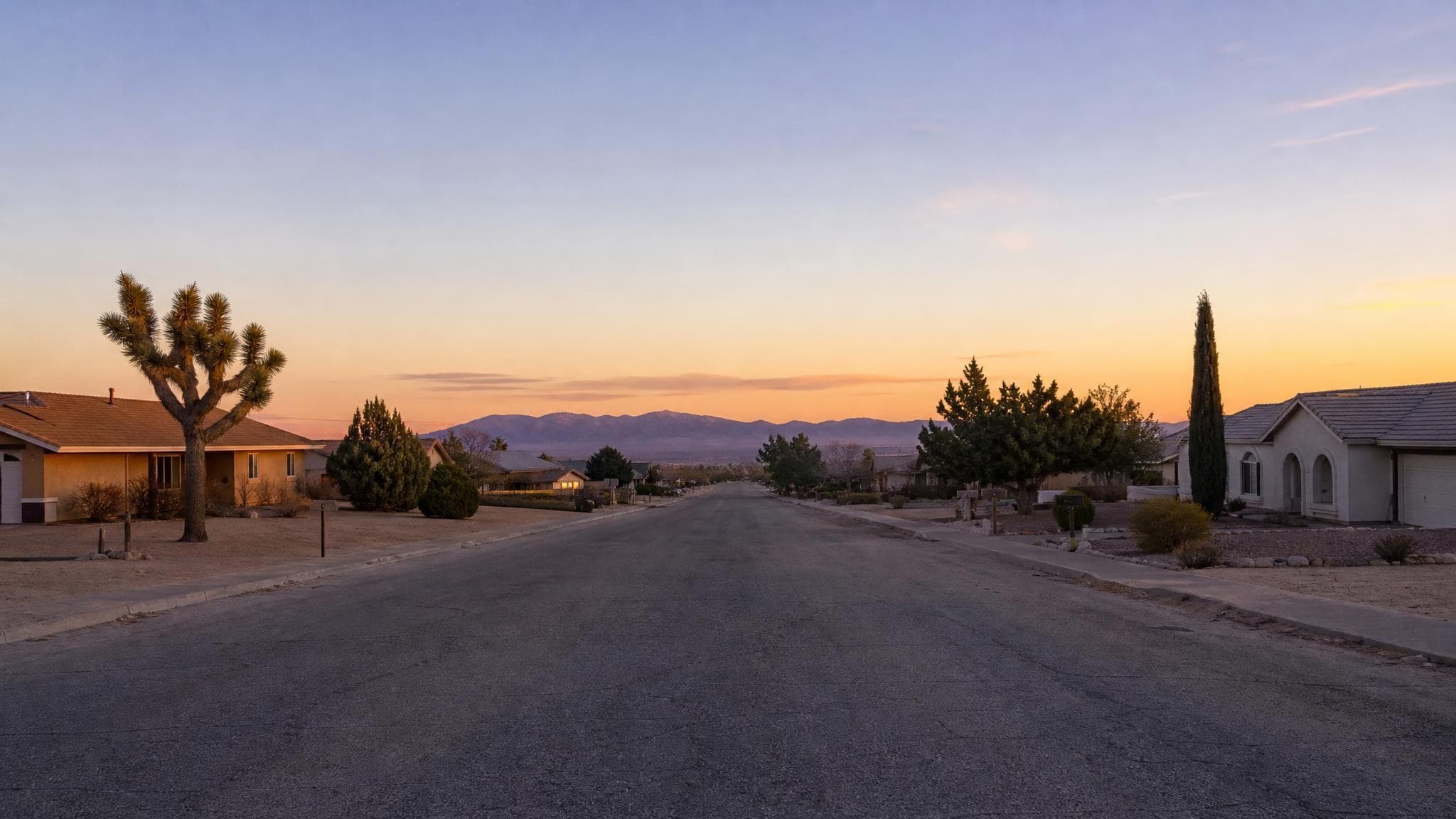A wide High Desert street of stucco homes in Victorville at sunset, with Joshua trees in the yards and mountain foothills purple on the horizon.